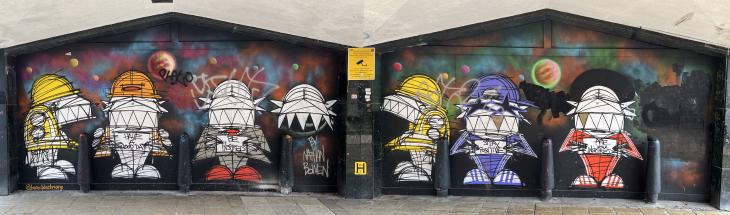 Graffiti Artwork stretching across two boarded up entrances to a shopping centre. Each entrance has 3 bollards across is creating 4 bays per entrance. In each of these bays is painted a cartoon graffiti figure with large teeth and a hat. Two are dressed as workmen in fluorescent jackets and hats. The rest are dressed in various outfits holding signs saying &lsquo;Have a good day&rsquo;, &lsquo;Be inspired&rsquo;, &lsquo;Say positive&rsquo; and &lsquo;Are you inspired?&rsquo;