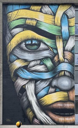 A painting on a brick facia of a building. It&rsquo;s the right half of a face made of intertwined ribbons of white, yellow, green, blue and red, that could resemble the muscles of the face, on a black background. The face looks friendly and a cityscape is visible in the reflection it its eye.