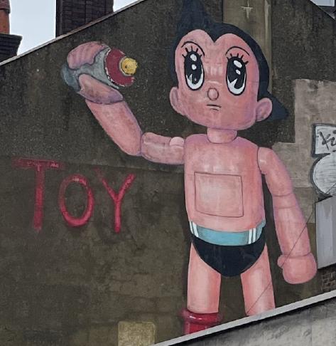 A graffiti painting on a wall above a parade of shops. The painting is of the cartoon character Astroboy, a skin coloured robotic character dressed just in underpants and red boots, holding a spray paint can in a defiant pose next to the spray-painted word &lsquo;Toy&rsquo;