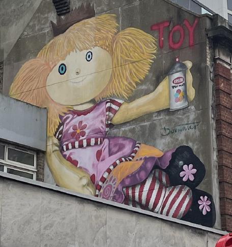 A graffiti painting on a wall above a parade of shops. The painting is of a ragdoll in a purple flowery dress holding a can of spray paint next to the spray-painted word &lsquo;Toy&rsquo;