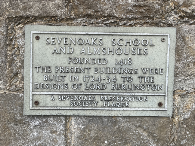 A grey plaque on a wall reading: Sevenoaks School and Almshouses. Founded 1418. The present buildings were built in 1724-34 to the designs of Lord Burlington. (A sevenoaks preservation society plaque)