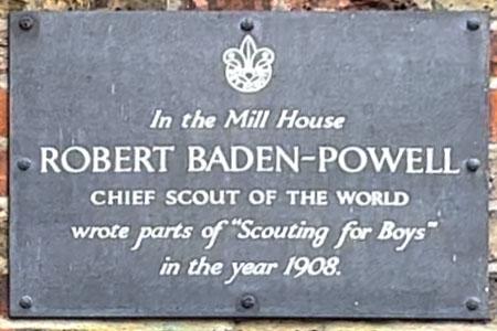 A dark grey metal plaque attached to a brick wall. It reads: &lsquo;In the Mill House Robert Baden-Powell chief scout of the world wrote parts of &ldquo;Scouting for Boys&rdquo; in the year 1908.&rsquo;